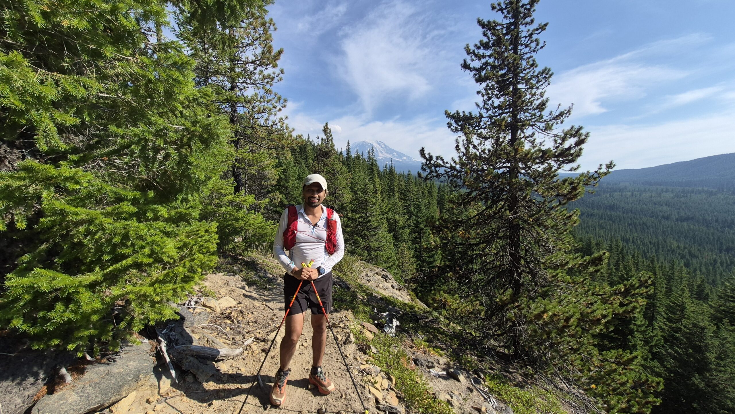 A trail runner with poles on a forest ridge, snow-capped peak behind